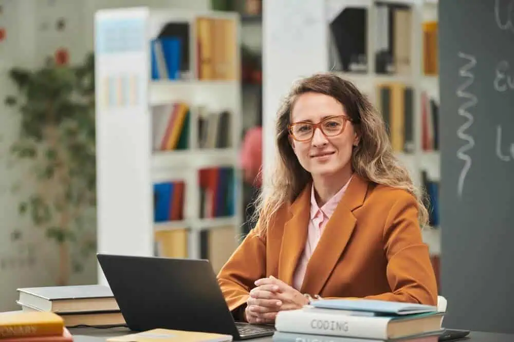 Female teacher using a laptop in a classroom setting with books and a chalkboard.