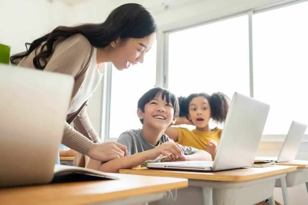 Teacher assisting students during online learning session in a classroom setting.