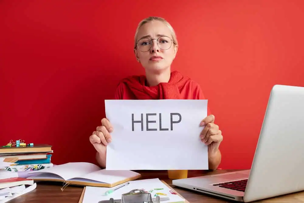 Help sign held by a distressed woman at a cluttered desk with a laptop and books, emphasizing the ne.