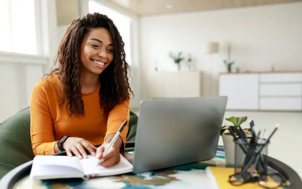 Smiling woman participating in an online learning session with a laptop and notebook.