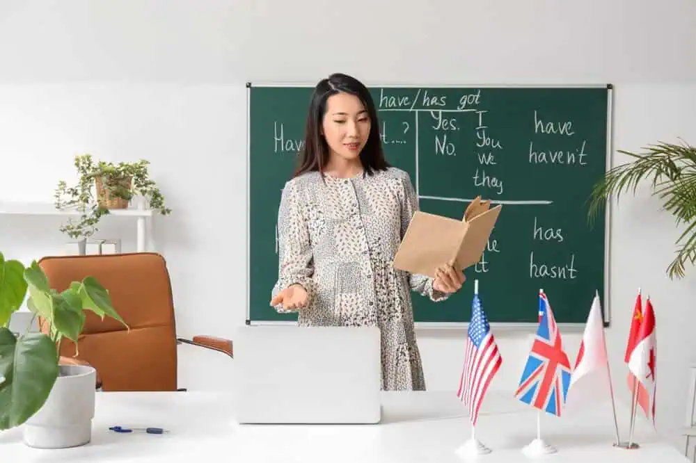 Female teacher conducting online class with flags and blackboard in background.