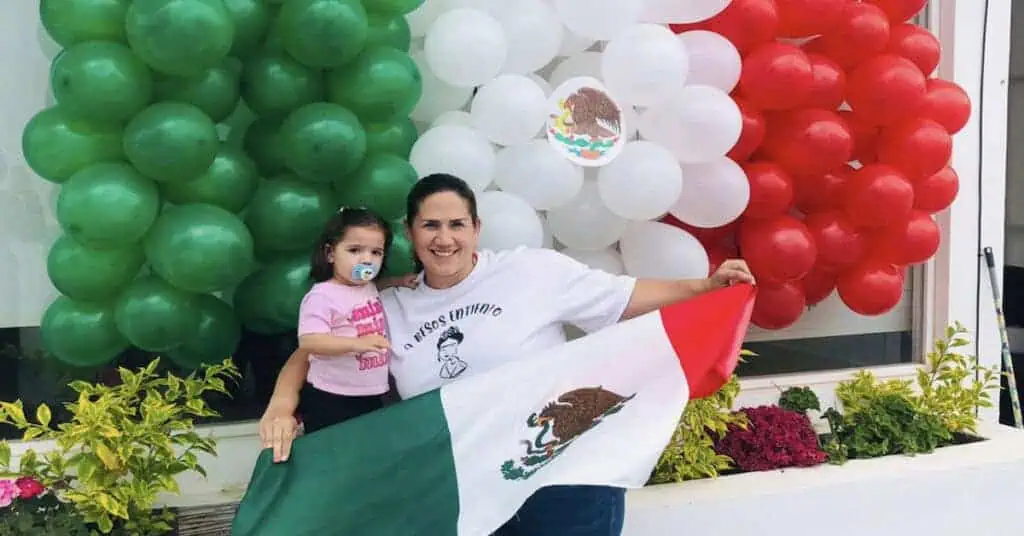 Woman and child celebrating with Mexican flag and balloons in front of a decorated backdrop.