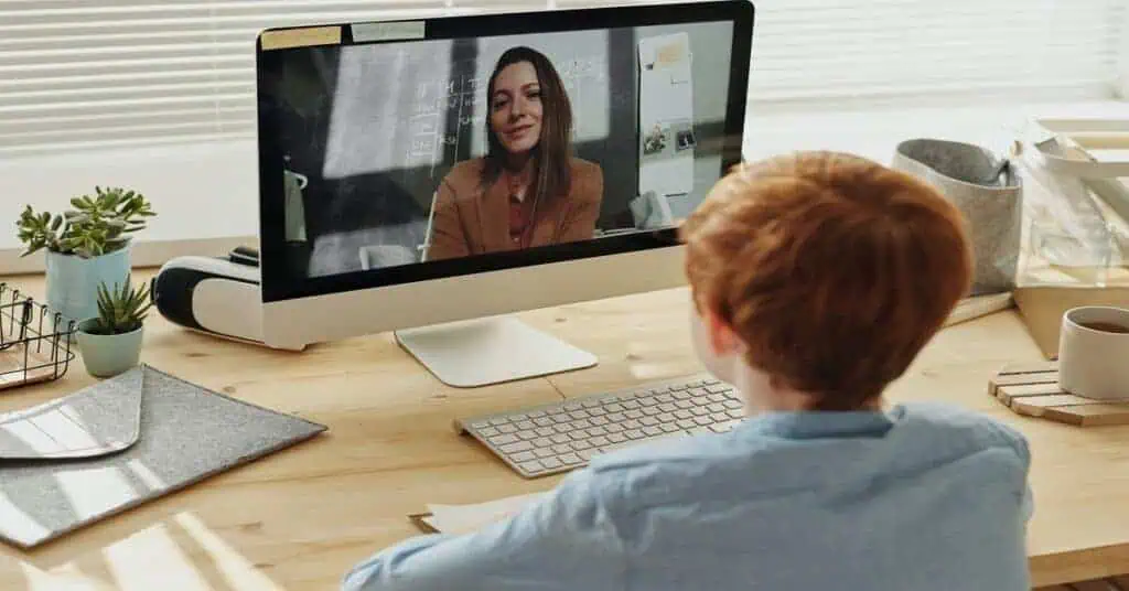 Student participating in a virtual classroom session with a teacher on a computer screen.