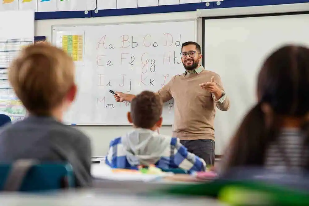 Teacher instructing students in a classroom with alphabet on whiteboard.