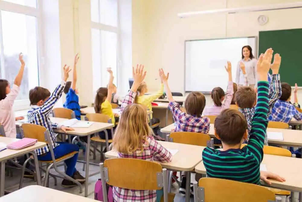 Engaged students raising hands in a classroom with a teacher at the blackboard.
