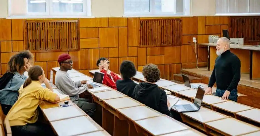 Multiple students sit in a college classroom with laptops open