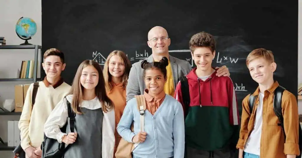 A live certified teacher stands smiling with six of his students in front of a chalkboard.