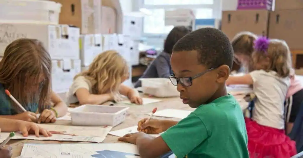 Young children sit at a long table doing schoolwork with papers and pencils.