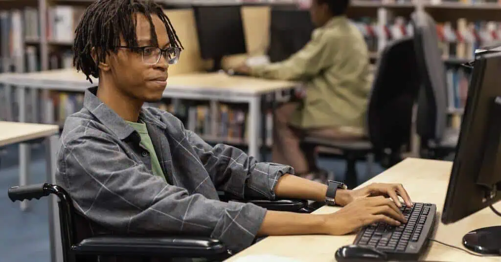 A young man in a wheelchair does schoolwork at a computer in a library.