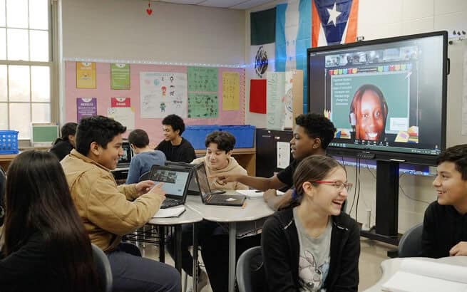 Students engaging in a virtual classroom with a teacher on a large screen.