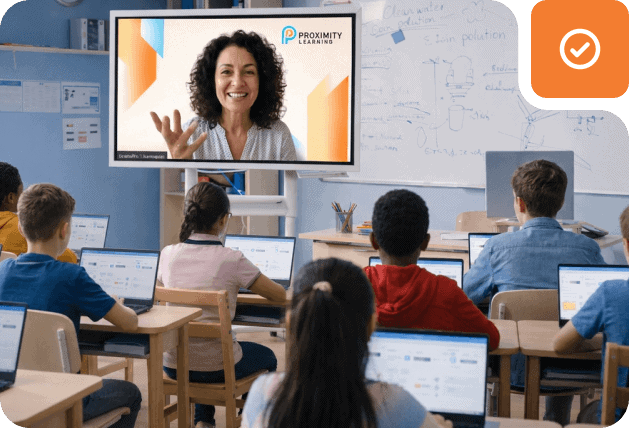 Students attending a virtual class with a teacher on a large screen in a classroom setting.