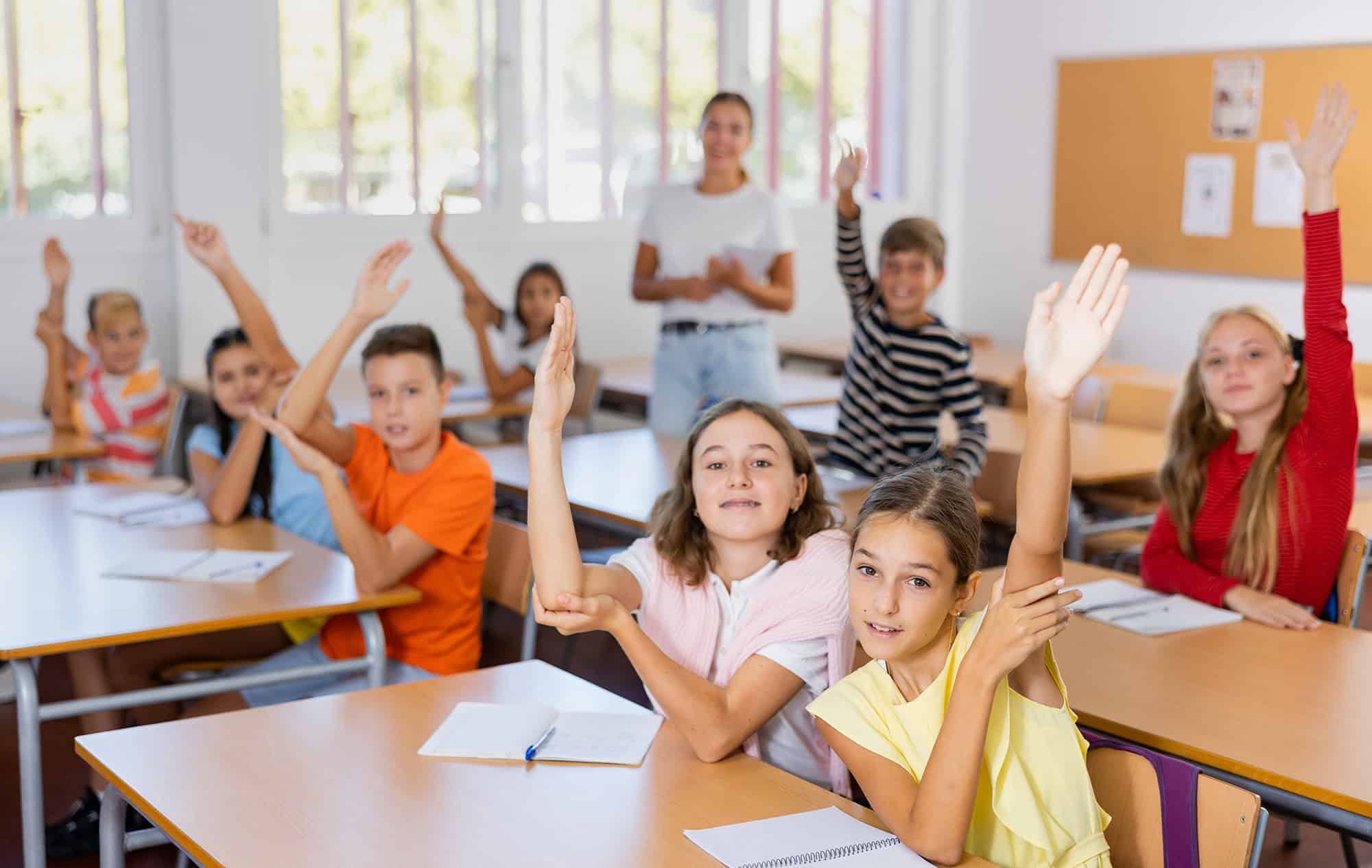 Students raising hands in classroom with teacher and bright windows.