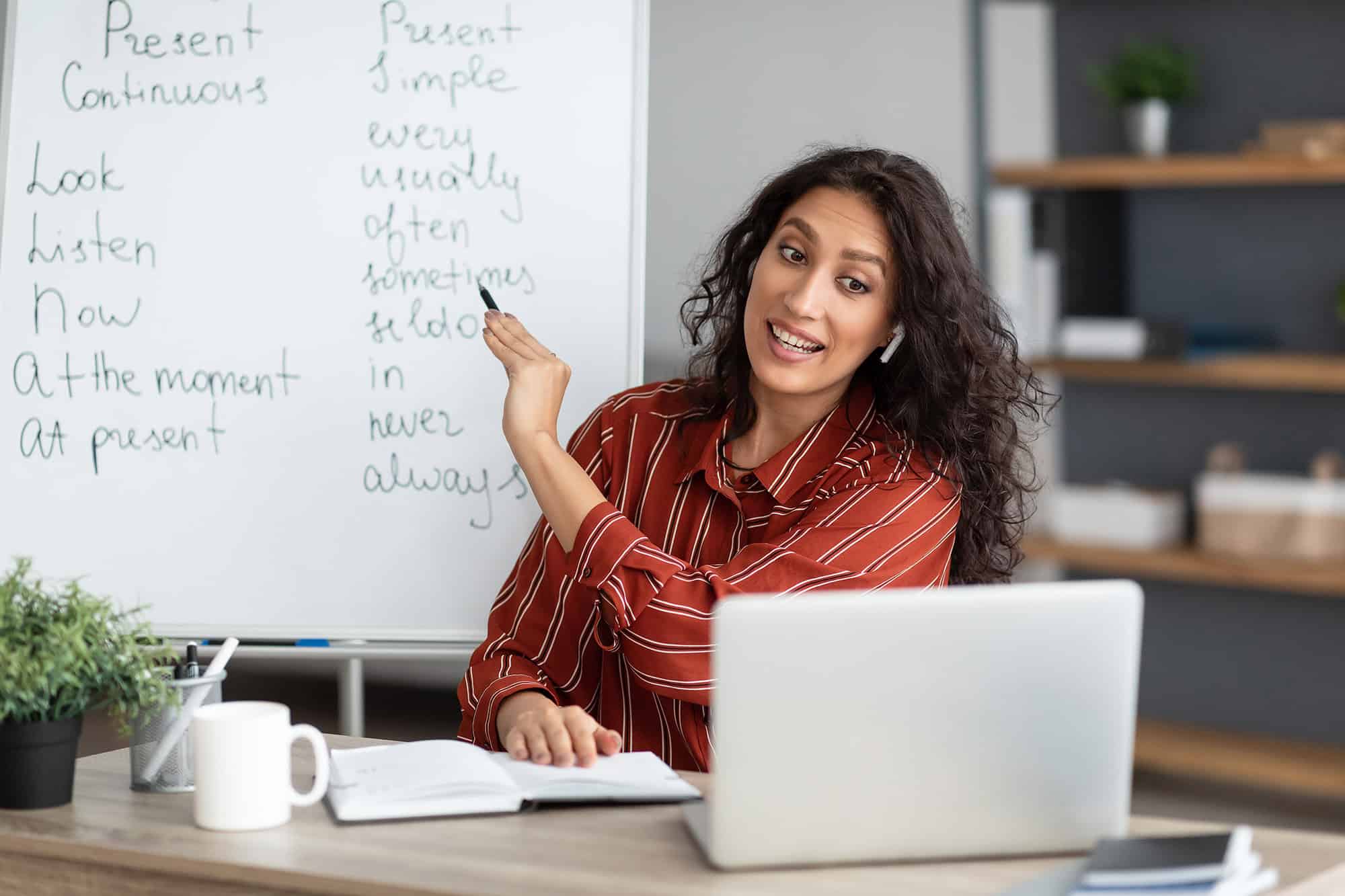 Female virtual teacher explaining present tense grammar on whiteboard.
